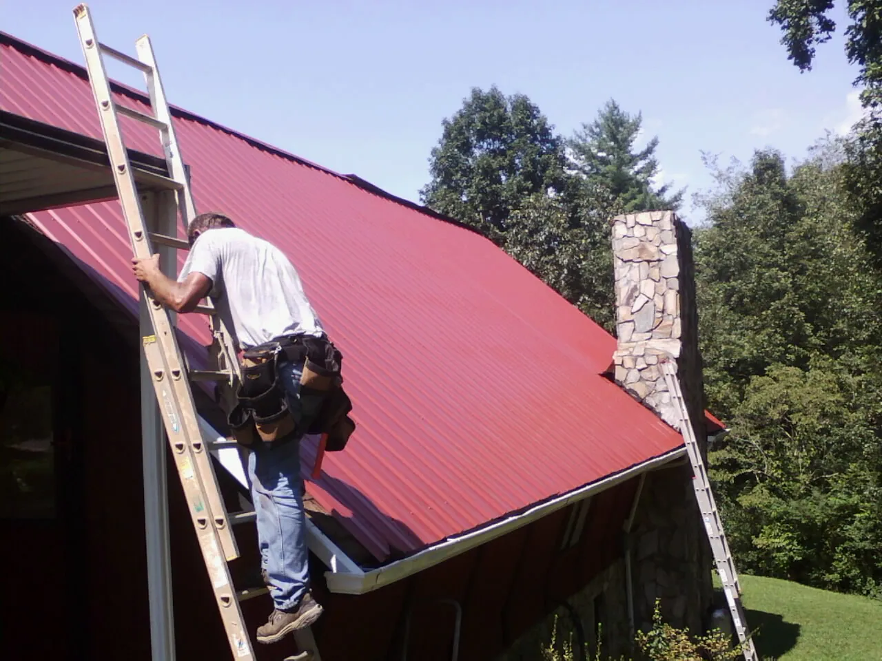 Roofer on a ladder beside a red metal roof and stone chimney — Soffit & Fascia Repair by Roofing Brothers in Harleysville